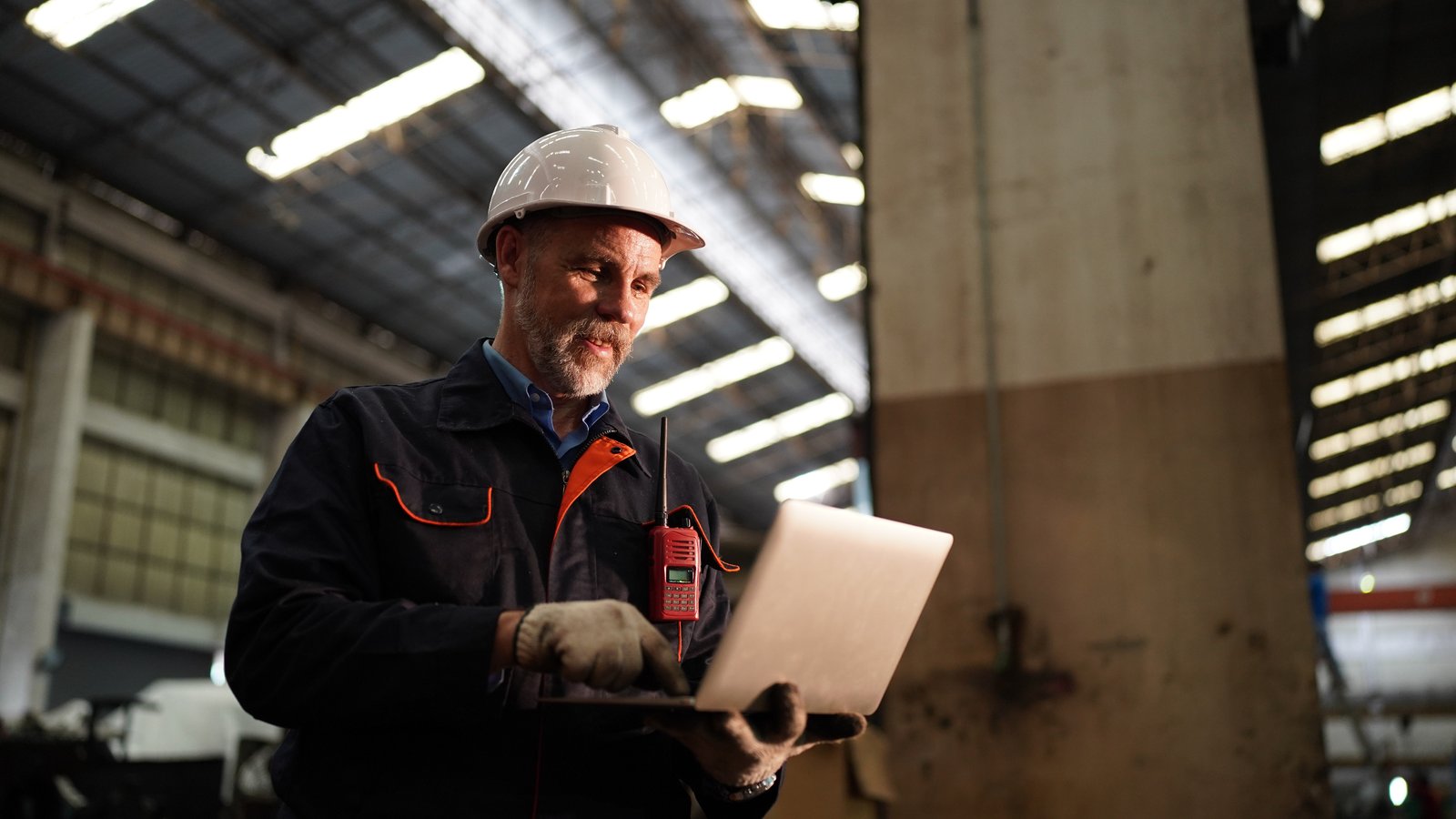 Worker in hard hat and safety gear holding a laptop in an industrial warehouse setting